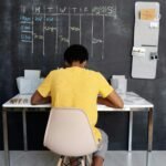 A teenager focuses on studying at a desk, with a blackboard displaying a schedule in the background.